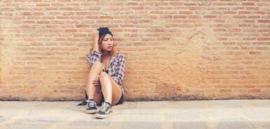 Jovem mulher triste sentada contra a parede de tijolos sozinha, ilustrando artigo sobre inferno astral na astrologia