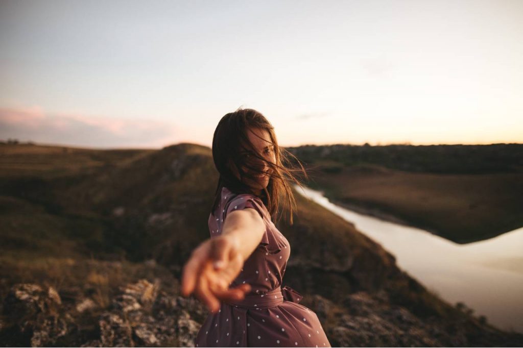 Imagem de uma mulher em uma viagem, por campos e rios, com uma mão direcionada para a lente que faz a fotografia, para ilustrar o artigo 'sagitário combina com qual signo?'