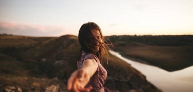 Imagem de uma mulher em uma viagem, por campos e rios, com uma mão direcionada para a lente que faz a fotografia, para ilustrar o artigo 'sagitário combina com qual signo?'