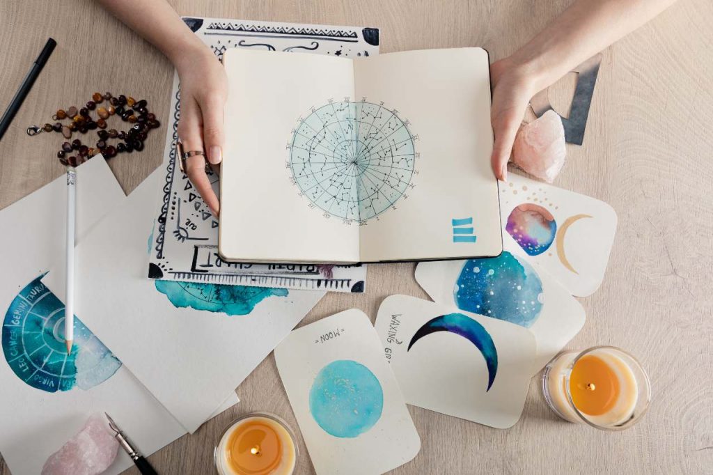 Vista superior de uma astróloga segurando caderno com desenhos em aquarela e signos do zodíaco em cartões na mesa, para ilustrar o horóscopo de hoje (16/10)