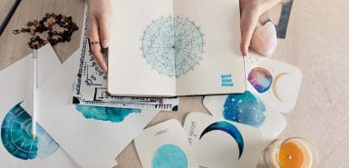 Vista superior de uma astróloga segurando caderno com desenhos em aquarela e signos do zodíaco em cartões na mesa, para ilustrar o horóscopo de hoje (16/10)