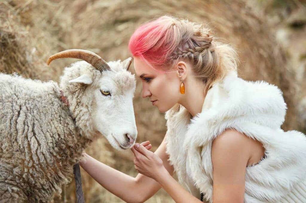 Mulher com roupa branca e cabelos coloridos encosta a cabeça na cabeça de um carneiro, símbolo do signo de Áries, para artigo sobre sobre aproveitar o sol em áries