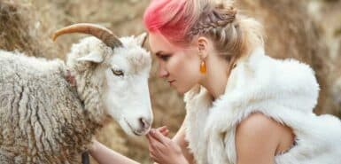 Mulher com roupa branca e cabelos coloridos encosta a cabeça na cabeça de um carneiro, símbolo do signo de Áries, para artigo sobre sobre aproveitar o sol em áries