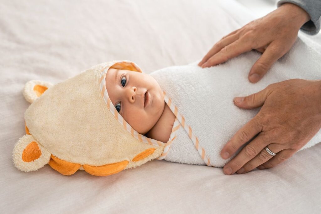 Bebê deitado e envolvido por uma toalha, mãos do pai.