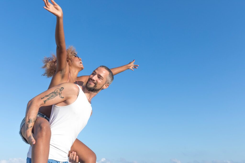 Homem carrega a namorada no estilo piggy back na praia no verão. Eles estão sorrindo