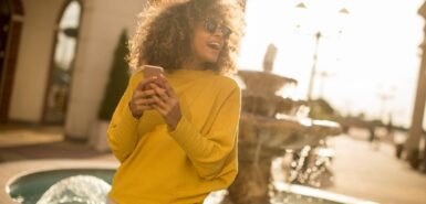 Jovem atraente com cabelo encaracolado usando seu celular em frente a uma fonte, com a luz do sol poente