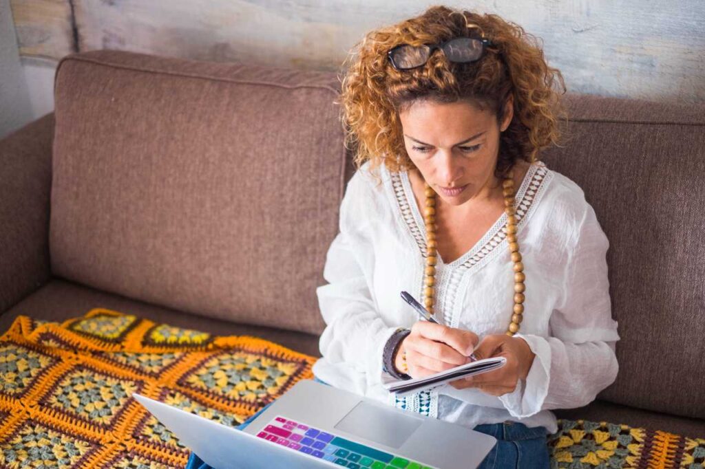 mulher de meia idade trabalhando em casa com um laptop enquanto está sentada no sofá. tomando notas e olhando para a área de trabalho do computador. internet office estilo de vida alternativo