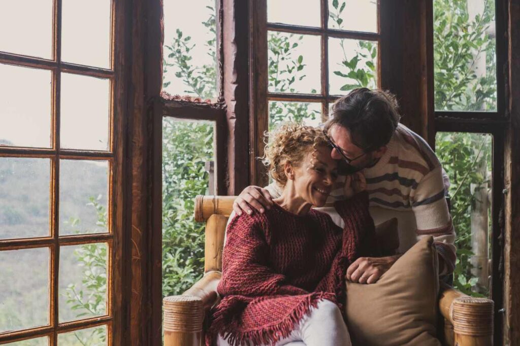Mulher sentada no sofá e homem a beija com ternura. Ao fundo, janelas que dão vista para a natureza verde