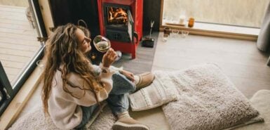 Jovem mulher sentada junto à lareira em um suéter branco, bebendo vinho em uma aconchegante cabana de madeira