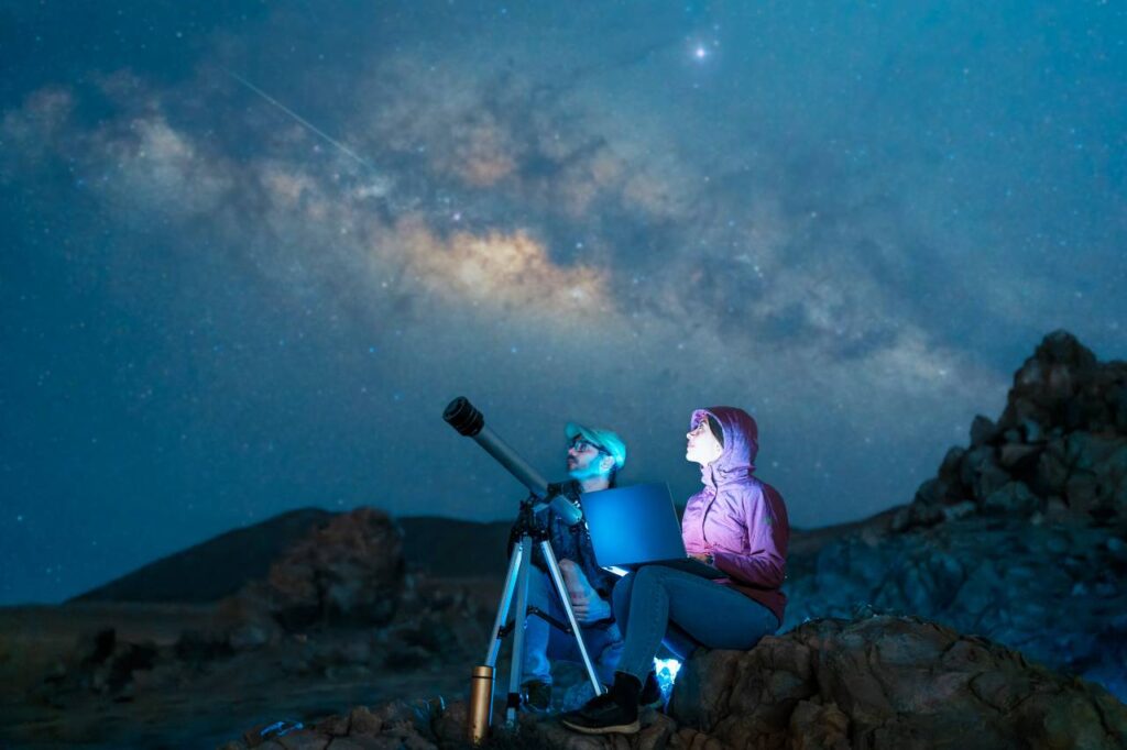 casal sentado no deserto observando as estrelas e a Via Láctea ao lado de um telescópio