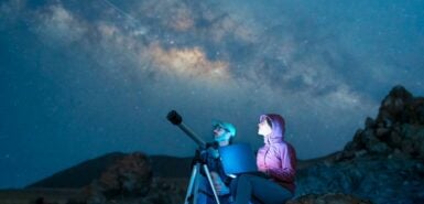 casal sentado no deserto observando as estrelas e a Via Láctea ao lado de um telescópio