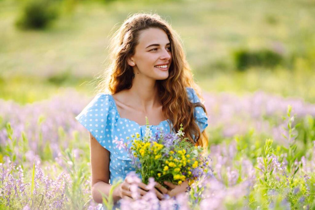 Retrato de uma mulher em um campo florido, segurando alguns ramos, com um vestido azul claro com babados e cabelos ondulados e soltos