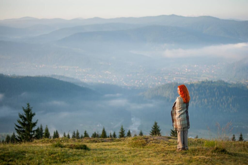 Mulher ruiva coberta com uma manta em pé em uma colina em bela paisagem montanhosa com neblina matinal sobre as montanhas e florestas