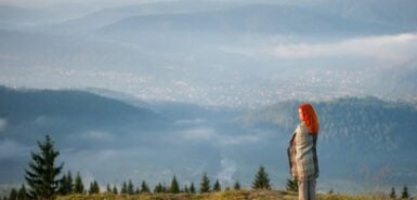 Mulher ruiva coberta com uma manta em pé em uma colina em bela paisagem montanhosa com neblina matinal sobre as montanhas e florestas