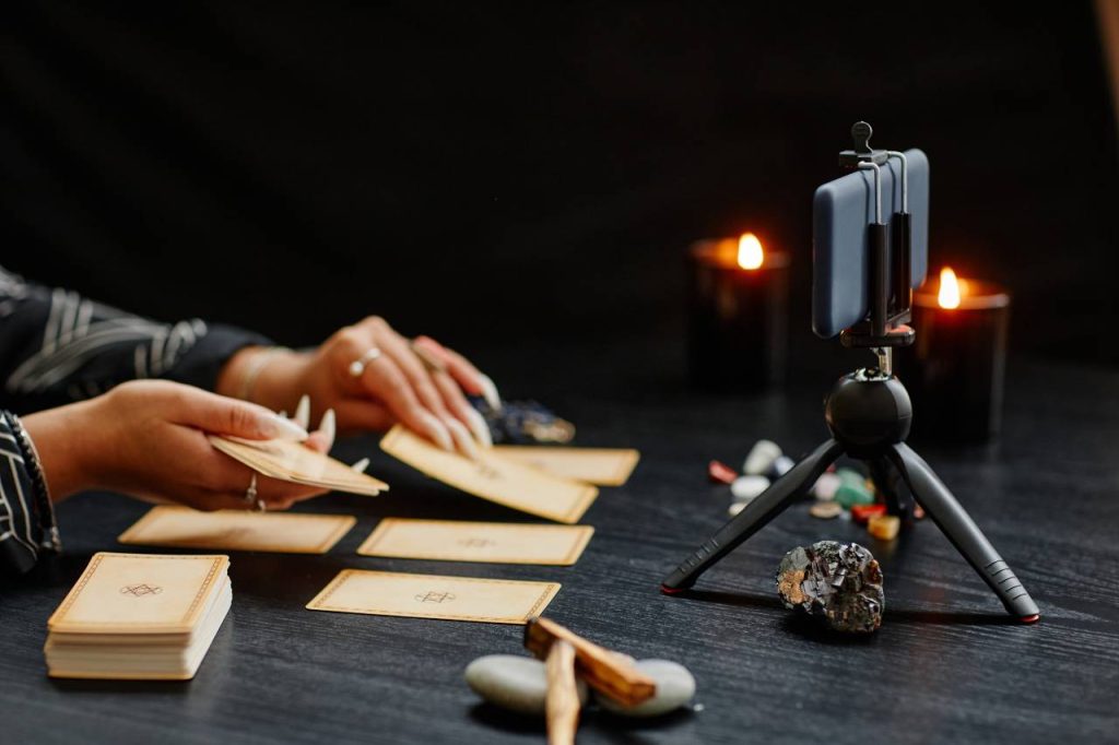 Mãos femininas fazem uma tiragem de tarot em frente a um celular em gravação, em uma leitura de tarot online