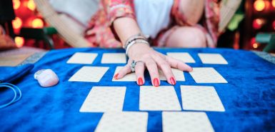 mão com unhas vermelhas e anel estendida por cima de cartas de tarot, em uma toalha de veludo azul